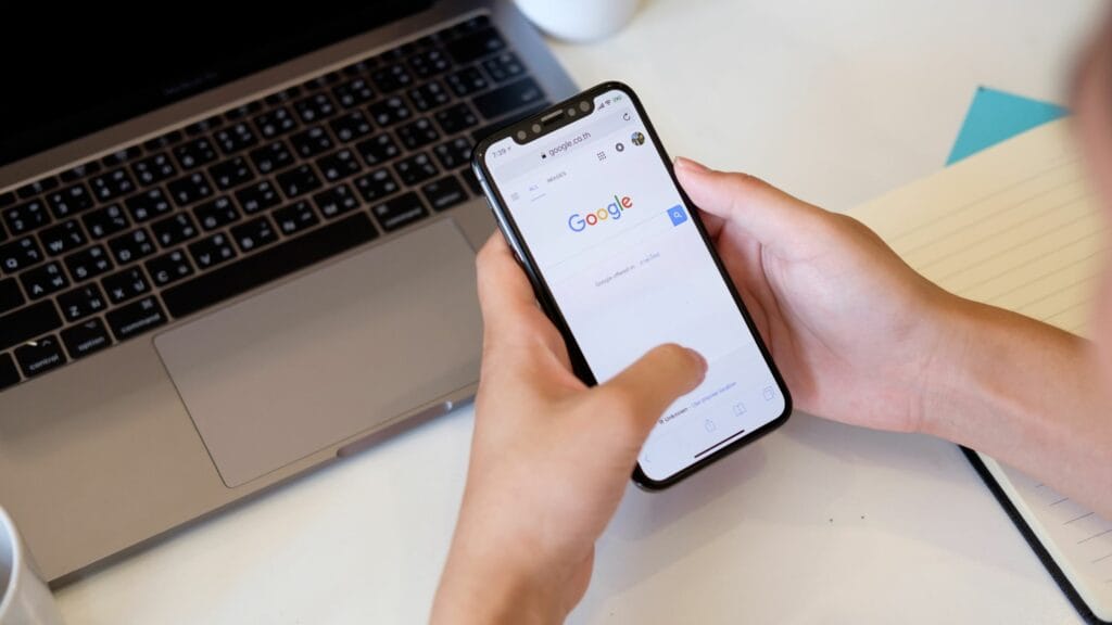 Close-up of hands holding a smartphone displaying the Google search homepage on a white desk with a laptop nearby.